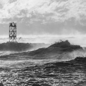 Op de afbeelding: Een zwart-witfoto van een hoge metalen constructie op een rotsachtige uitstulping in het midden van een stormachtige zee. De golven beuken tegen de rotsen en de wind zwiept de spatten omhoog.