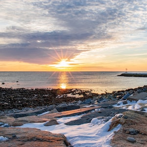 Op de afbeelding: Een schilderachtig kustlandschap bij zonsondergang. De zon, een heldere ster, weerspiegelt in het kalme water. Een rotsachtige kustlijn met sneeuwplekken leidt naar een stenen pier. De lucht is een mix van oranje, geel en blauw.