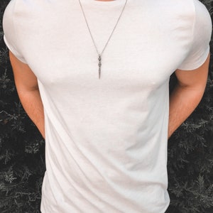 May include: A silver chain necklace with a silver dagger pendant. The necklace is worn by a person wearing a white t-shirt.
