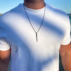 May include: A silver chain necklace with a silver dagger pendant. The pendant has a detailed design and is hanging from a thin chain.