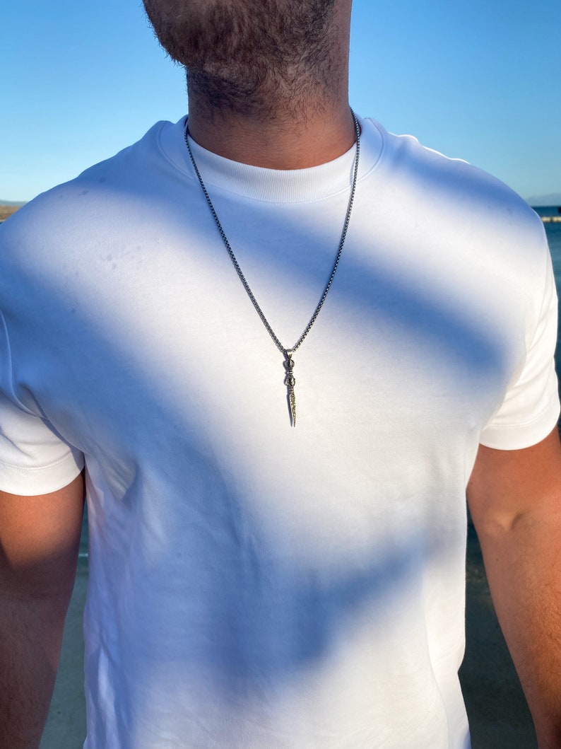 May include: A silver chain necklace with a silver dagger pendant. The pendant has a detailed design and is hanging from a thin chain.