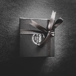 May include: A black gift box with a black ribbon tied in a bow. The box has a white sticker with the text "SPARK AT&Ouml;LYE" and "HANDMADE" in a circle.