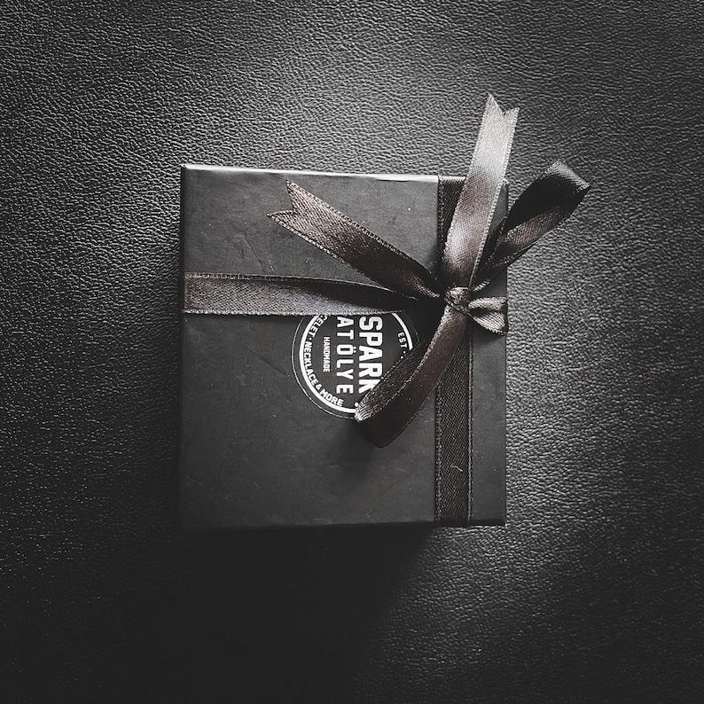 May include: A black gift box with a black ribbon tied in a bow. The box has a white sticker with the text "SPARK AT&Ouml;LYE" and "HANDMADE" in a circle.