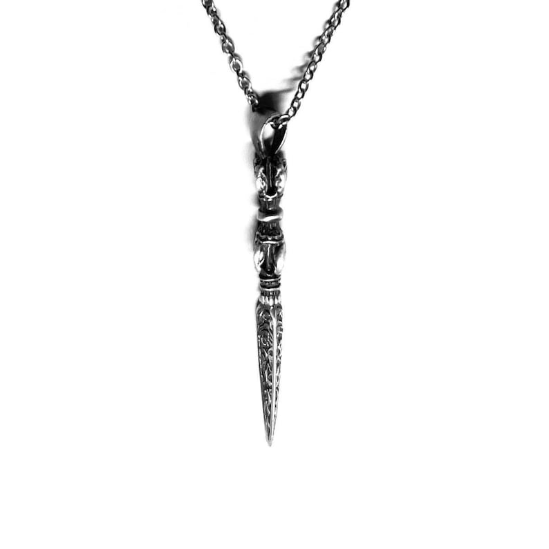 May include: A silver pendant necklace featuring a detailed dagger design. The dagger has a pointed tip and intricate carvings along its length.