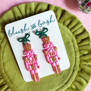 May include: A pair of dangle earrings featuring pink nutcracker figures with gold glitter accents, each topped with a small dark green bow. The earrings are displayed on a white card printed with the text blush and bash studio in dark blue.