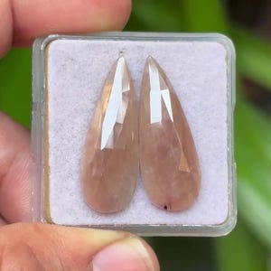 May include: Two faceted, teardrop-shaped gemstones in a clear plastic case. The gems have a peach-pink hue and are displayed against a white background. The gems are held by a hand.