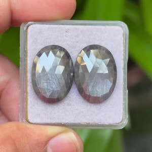 May include: Two oval-cut gemstones in a clear plastic case. The stones are a smoky grey colour with internal veining and a brilliant, reflective surface. The gemstones are displayed on a light pink background.