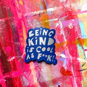 Peut inclure: Un autocollant découpé avec un fond bleu foncé et des lettres à paillettes argentées qui indiquent "BEING KIND IS COOL AS F**K!" dans une police grasse et empilée. L'autocollant a une forme organique irrégulière et une finition scintillante.