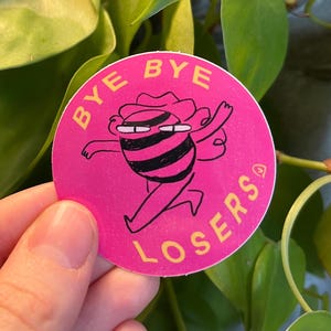 Könnte beinhalten: Ein runder rosa Aufkleber mit einer schwarz-weißen Comicfigur, die weggeht, mit dem Text "BYE BYE LOSERS" in Gelb.