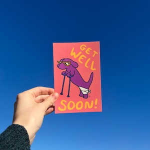 Peut inclure: Carte de vœux rose avec un chien de dessin animé violet portant des lunettes de soleil et des béquilles. Le texte jaune dit « Get Well Soon! ». L'image est prise contre un ciel bleu clair, créant une ambiance joyeuse.