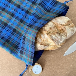 Könnte beinhalten: Ein Brot in einem blau, grün und schwarz karierten Brotbeutel. Ein gezahntes Messer liegt neben dem Brot. Der Beutel hat ein kleines Etikett. Der Brotbeutel ist aus Stoff.