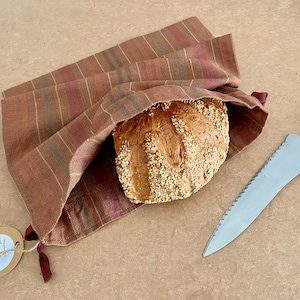 Könnte beinhalten: Ein Brot mit Sesamkörnern ist in einem braunen und roten gestreiften Stoffbeutel verpackt. Ein Brotmesser liegt neben dem Beutel auf dem Tisch.
