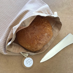 Könnte beinhalten: Ein Brotlaib in einer beigefarbenen Leinentasche für Brot mit einem braunen Papieretikett mit der Aufschrift "Kühlschrank" und Waschhinweisen. Ein gezacktes Brotmesser liegt neben der Tasche auf dem Tisch.
