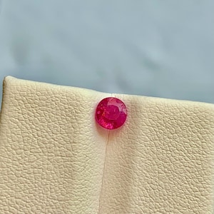 Puede incluir: Una gema de rubí redonda y facetada. El rubí es de un tono rojo vibrante y está colocado sobre una superficie texturizada de color blanquecino. El fondo es de un azul claro y suave. Esta imagen muestra una piedra preciosa.