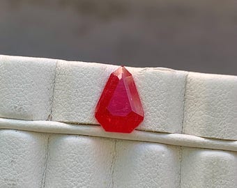 May include: A faceted, red gemstone with a pointed top and a geometric shape. The gem is set against a white, textured surface. The gemstone's color is a vibrant, translucent red.