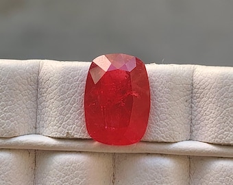 May include: A faceted, oval-shaped, red gemstone. The gem has a polished surface with multiple facets that reflect light. The gemstone is set against a white, textured background.