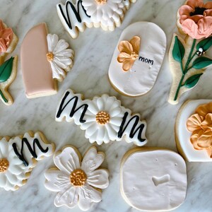 May include: Assortment of decorated sugar cookies with floral and "mom" designs. Cookies feature white icing, peach and orange flowers, green leaves, and black script. The cookies are arranged on a marble surface.