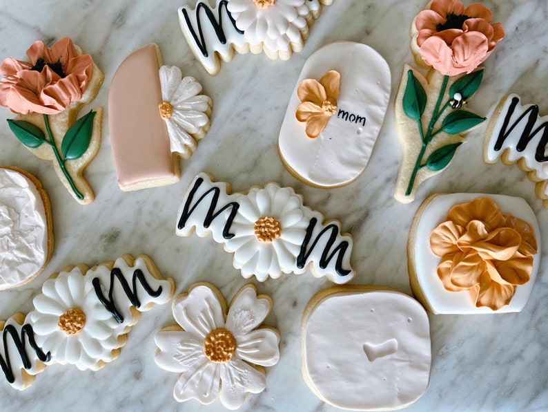 May include: Assortment of decorated sugar cookies with floral and "mom" designs. Cookies feature white icing, peach and orange flowers, green leaves, and black script. The cookies are arranged on a marble surface.