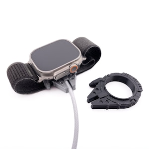 May include: A silver smartwatch with a black band is attached to a 3D printed stand. A black 3D printed object shaped like a spaceship is in the background.