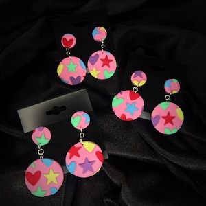 May include: Three pairs of dangle earrings with pink, red, yellow, green, and blue hearts and stars on a pink background. The earrings are on black card stock.