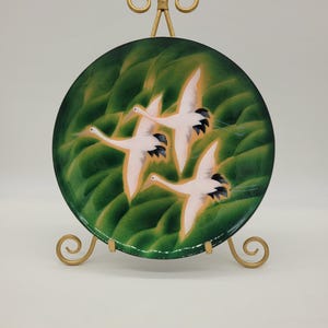 May include: A decorative plate featuring three white birds in flight against a green background. The plate is displayed on a gold-colored metal stand. The birds have black wingtips and are painted with fine detail.