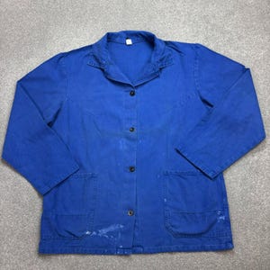Può includere: Giacca da lavoro blu vintage con collo, tre bottoni neri e due ampie tasche frontali. La giacca ha maniche lunghe e mostra segni di usura, con alcuni segni di vernice bianca.