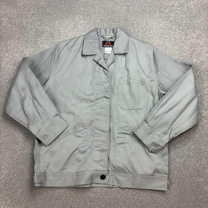 May include: A light grey long-sleeve work jacket featuring a pointed collar, a single chest pocket on the left side, and buttoned cuffs. The garment includes a front button closure and a black label at the inner neckline with the text 100.
