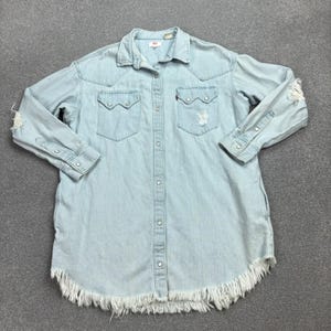 May include: Light blue denim shirt with long sleeves, two chest pockets, and a button-down front. The shirt has distressed details on the sleeves and a fringed hem. A small red Levi's tag is visible on the chest pocket.