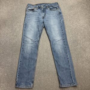 May include: Light blue denim jeans with a classic five-pocket design. The jeans have a straight leg cut and a button and zipper closure. The denim has a faded wash effect, giving them a worn-in look. The jeans are laid flat on a gray surface.
