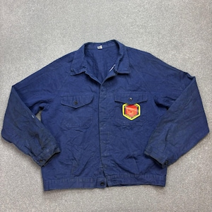 May include: A navy blue work jacket with two chest pockets and a button-up front. The jacket has a colourful patch on the chest. The jacket has a collar and long sleeves. The fabric appears worn, suggesting it is a well-used garment.