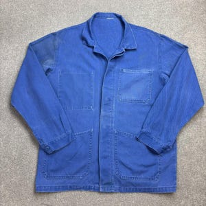 May include: A long-sleeved blue work jacket featuring a classic collar, a button-down front, and four large patch pockets on the front. The garment is constructed from a durable fabric with visible stitching details and buttoned cuffs on the sleeves.
