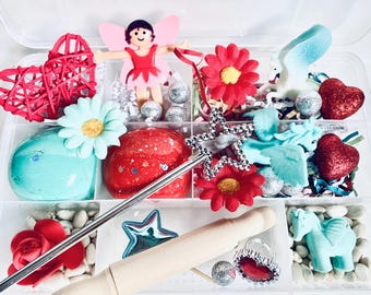 May include: A clear plastic storage box filled with various craft supplies for pretend play. The box includes red and blue play dough, a wooden rolling pin, a silver wand, a silver star, a red rose, white beans, a red heart, a silver ring, a blue unicorn, a silver fairy, and red and white flowers.