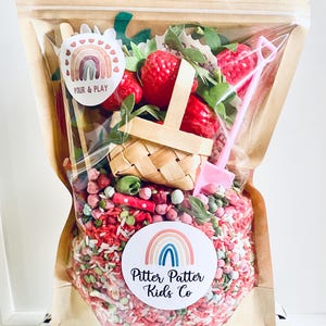 Puede incluir: Una bolsa de plástico transparente llena de artículos de juego sensoriales. La bolsa contiene arroz rojo y verde, fresas de juguete, una cesta tejida y una pala rosa. La bolsa tiene dos pegatinas con diseños de arcoíris y el texto "Pour & Play" y "Pitter Patter Kids Co."