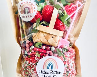 May include: A clear plastic bag filled with sensory play items. The bag contains red and green rice, toy strawberries, a woven basket, and a pink shovel. The bag has two stickers with rainbow designs and the text "Pour & Play" and "Pitter Patter Kids Co."