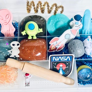 May include: A clear plastic storage box filled with space-themed toys and play dough. The box contains a green alien, a pink alien, a rocket, a spaceship, a moon, a rolling pin, a NASA sticker, and a blue and silver glitter ball.