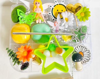 May include: A clear plastic container filled with colorful craft supplies. Includes a green butterfly, a yellow fairy figurine, a smiling sun sticker, a gold heart, and a wooden rolling pin. Also features a green star-shaped cutter and various floral and pom-pom accents.