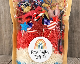 Puede incluir: Una bolsa transparente llena de capas de arroz teñido de rojo, blanco y azul, con figuras de dinosaurios de plástico, estrellas brillantes, banderas estadounidenses en miniatura y una pala negra, con el logo de arcoíris Pitter Patter Kids Co.