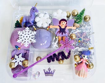 May include: A clear plastic container filled with fairy-themed toys and craft supplies. Includes a purple unicorn, fairy figurines, snowflake ornaments, a wand, and glittery gold baubles. The container is filled with purple playdough.