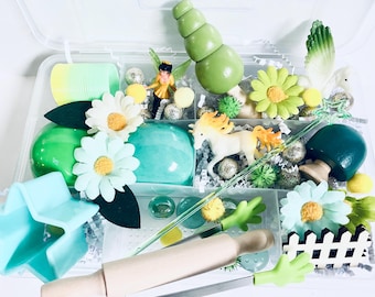 May include: A green and white fairy garden playset in a clear plastic storage container. The set includes a green play dough, a unicorn figurine, a fairy figurine, a rolling pin, a green mushroom, a green tree, a white picket fence, a star-shaped cookie cutter, and other small accessories.