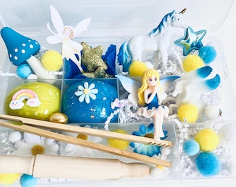 May include: A clear plastic storage box filled with blue and yellow play dough, a unicorn, a fairy, a mushroom, and other small toys. The box is filled with white shredded paper.