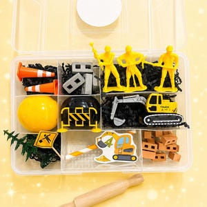 May include: A clear plastic container filled with construction-themed toys. Includes yellow construction worker figures, orange traffic cones, a yellow excavator, a yellow bulldozer, and miniature bricks. A wooden rolling pin is in the foreground.
