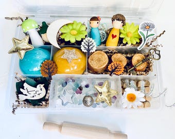 May include: A clear plastic container filled with various craft supplies. Includes wooden shapes like stars, a moon, and a tree, along with painted figurines, flowers, and a rolling pin. The color palette features blues, yellows, greens, and gold accents.