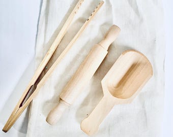 May include: A set of three wooden kitchen tools in a white cloth bag. The tools include a rolling pin, a scoop, and a pair of tongs.