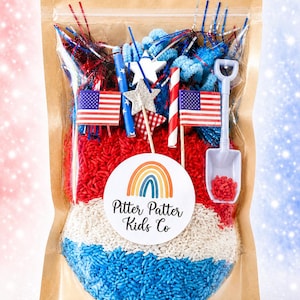 May include: A clear plastic pouch with a kraft paper border containing red, white, and blue dyed rice. Inside are two miniature American flags, a white plastic shovel, star-shaped cutouts, red and white striped straws, and blue pipe cleaners. A circular label features a rainbow and the text Pitter Patter Kids Co.