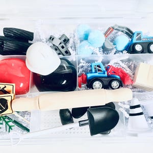 May include: A clear plastic storage container with various construction-themed toys, including a toy truck, a toy excavator, a toy hard hat, a toy shovel, and a toy wooden block. The container is filled with white shredded paper.