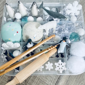 Puede incluir: Un contenedor de almacenamiento de plástico transparente lleno de varios artículos para una caja sensorial, que incluye plastilina blanca y azul, pompones blancos, copos de nieve blancos, un oso polar, pingüinos, una ballena y un rodillo de madera.