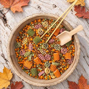 May include: A round wooden bowl filled with a mixture of dried orange lentils, brown rice, green split peas, and various pasta shapes including pumpkins, leaves, and ruffled rectangles, accompanied by a small wooden scoop and two wooden tongs.