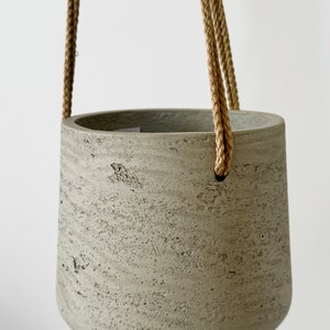 May include: A gray concrete hanging planter with a textured surface. The cylindrical pot is suspended by a thick, braided rope. The planter has a rounded bottom and a wide opening, suitable for plants.
