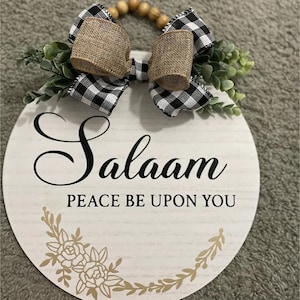 May include: A white round wooden sign with the words "Salaam Peace Be Upon You" in black lettering. The sign is decorated with a black and white checkered bow with burlap accents and green foliage.