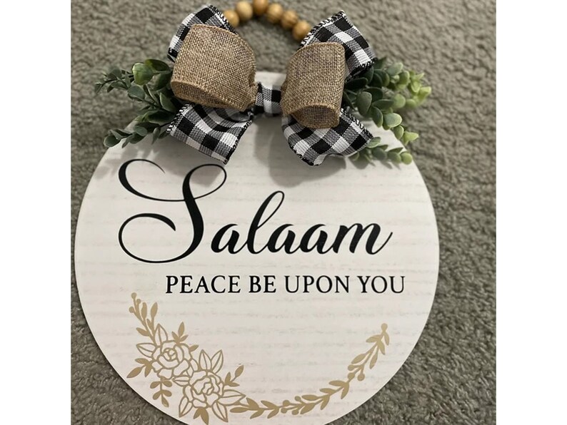 May include: A white round wooden sign with the words "Salaam Peace Be Upon You" in black lettering. The sign is decorated with a black and white checkered bow with burlap accents and green foliage.